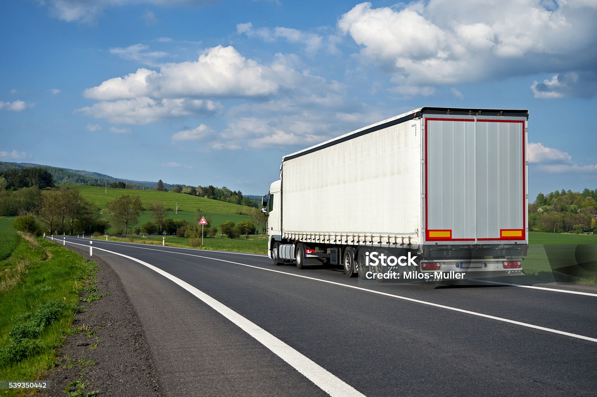 White truck departing on an asphalt road meander trough the green fields. Warning traffic sign Bend to right. Wooded mountains in the background. Sunny spring day with blue skies and white clouds.